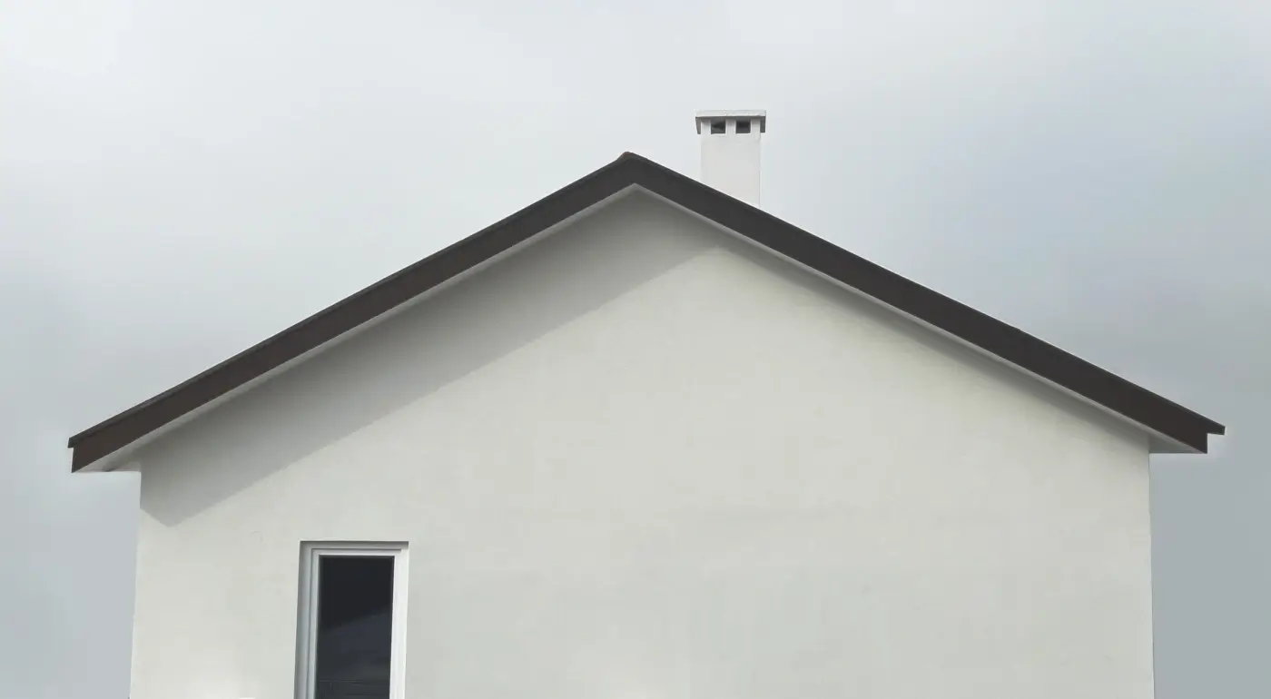 Plain white house with dark brown pointed roof line, a single dark window, and a white chimney against a flat gray sky.