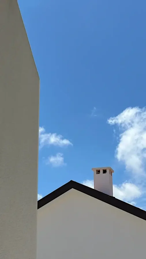 Plain white house with pointed roof and white chimney, partially blocked by a wall. The sky is deep blue with sparse clouds.